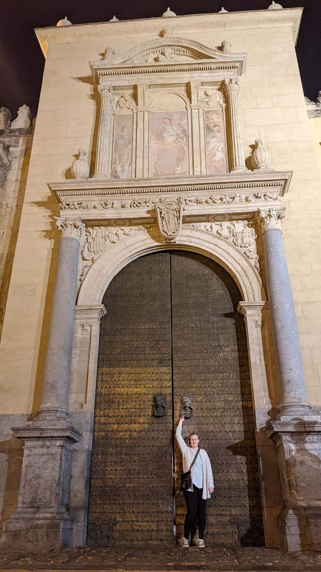 Anne stands at the foot of an enormous ornate cathedral door at night, dwarfed by the carved Renaissance facade above her