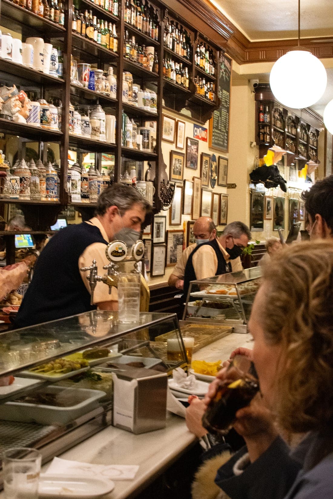 An old Madrid tavern with floor-to-ceiling bottles, framed press clippings on the walls, and bartenders behind a polished counter